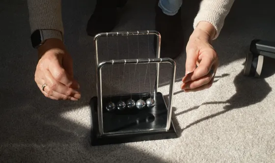 Twee handen rond een Newton-pendel, in scherp zonlicht